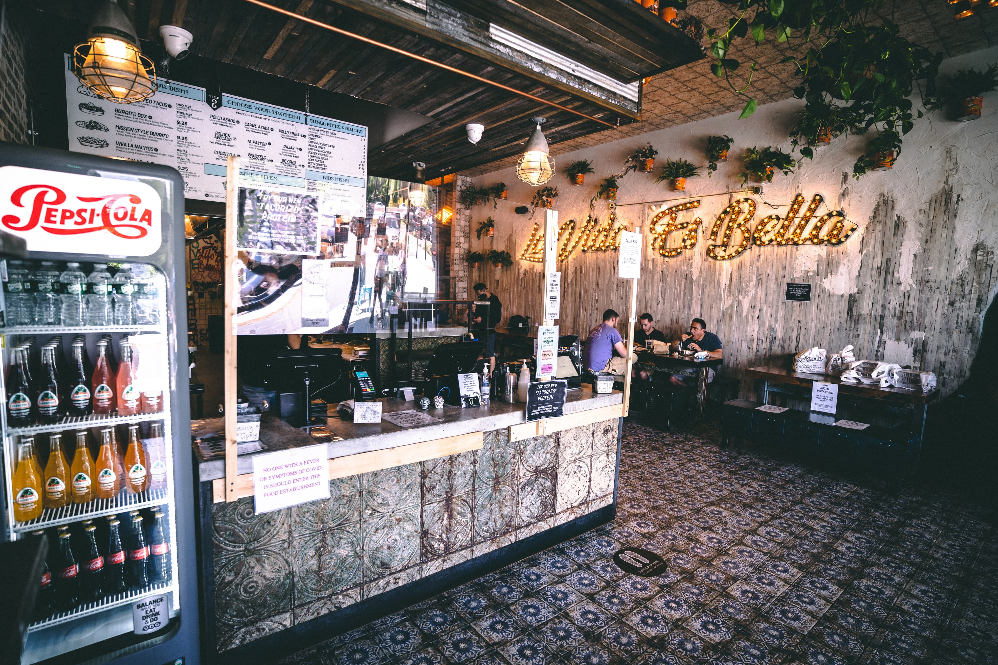 A wide shot of the ordering counter and beverage cooler, with the restaurant's menu board overhead. In the background, a lit-up marquee sign on a rustic wall reads "La Vida Es Bella" near patrons seated at tables.