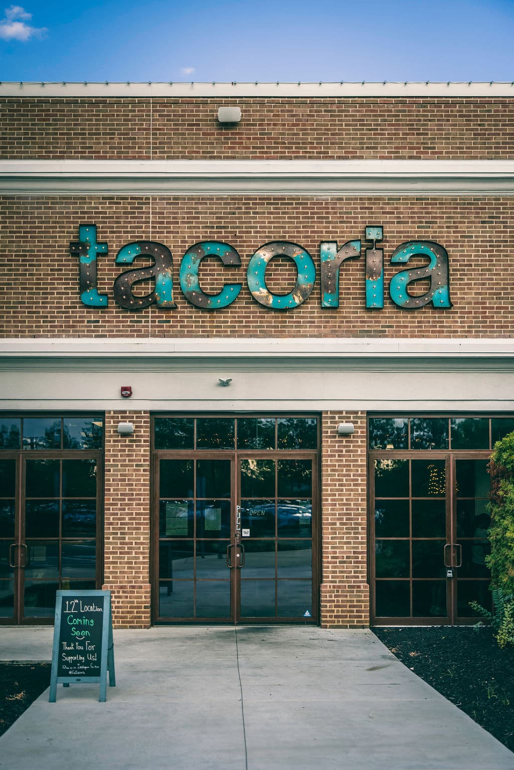 A straight-on shot of the brick exterior of Tacoria. Above the glass double doors, large, industrial-style letters spell out the restaurant’s name in a weathered teal patina. To the left of the entrance, a small chalkboard sign reads, "12th Location Coming Soon – Thank You For Supporting Us!"