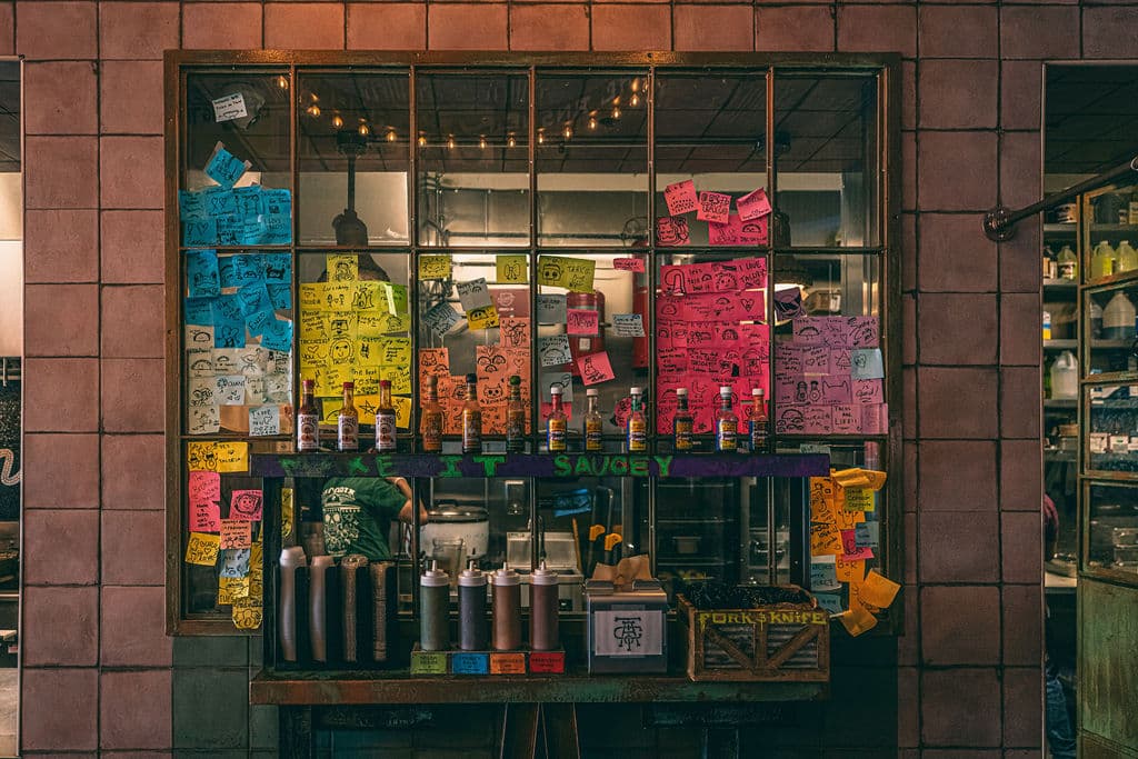 A sauce station set against a pink tiled wall. A large window frame is covered in colorful sticky notes with drawings and messages from customers. Below the window, several bottles of hot sauce and squeeze bottles are lined up on a shelf labeled "TAKE IT SAUCEY".