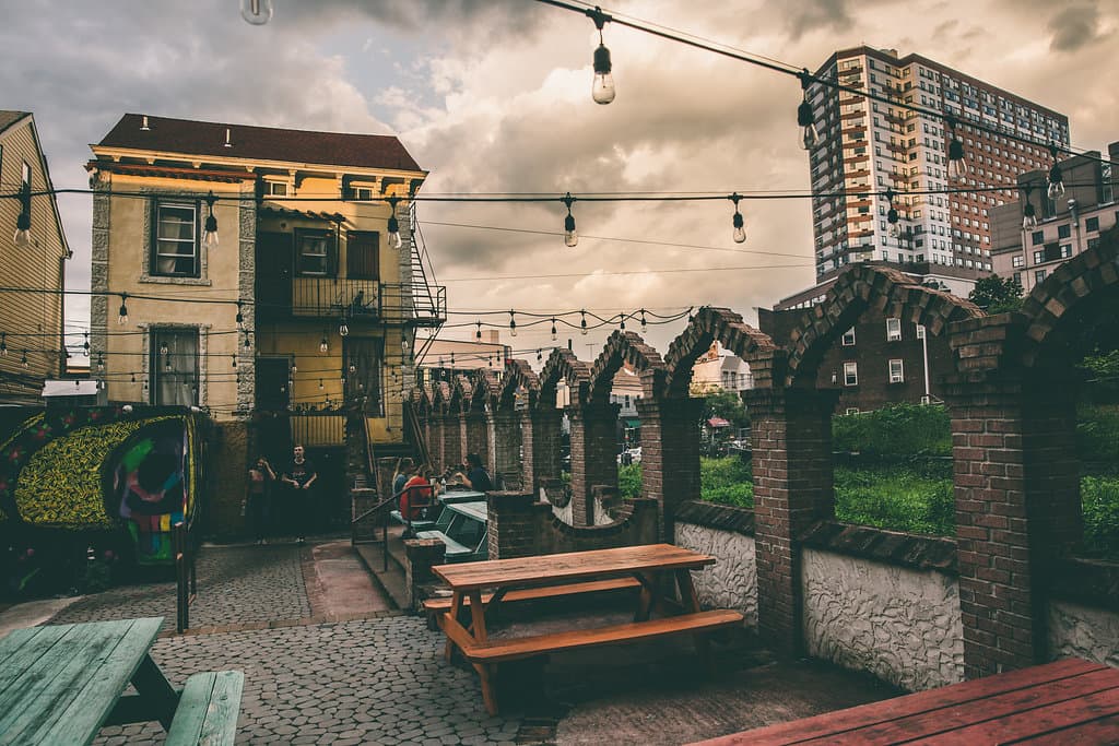 An outdoor patio area with wooden picnic tables and cobblestone flooring under a cloudy sky. String lights hang across the space, which is bordered by a brick wall with decorative arches and a view of a tall city building in the distance.