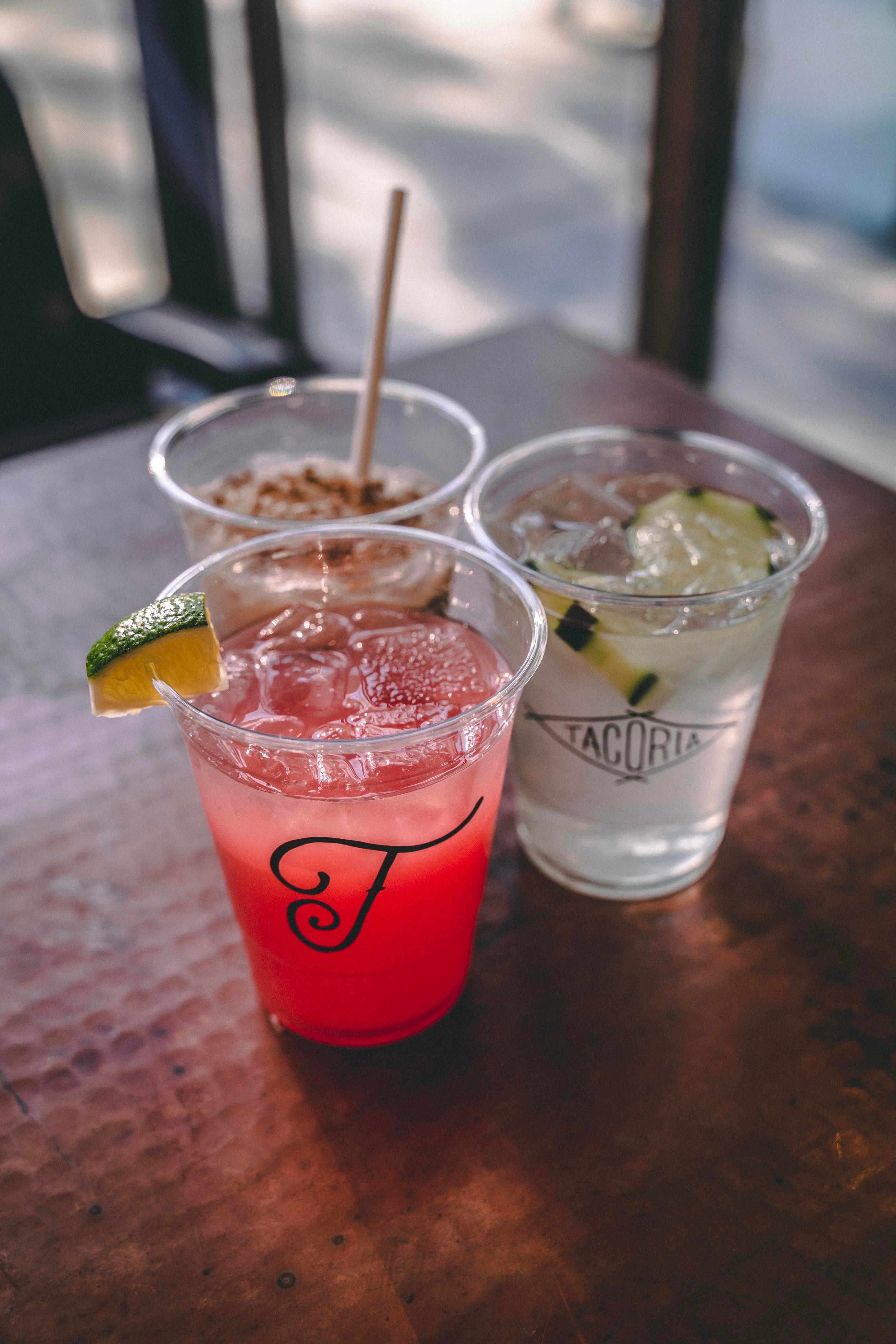 Three different colorful drinks in clear plastic cups sitting on a hammered copper tabletop. One is a bright red juice with a lime wedge, one is clear with cucumber slices and a "Tacoria" logo, and the third is a tan-colored horchata with a straw.